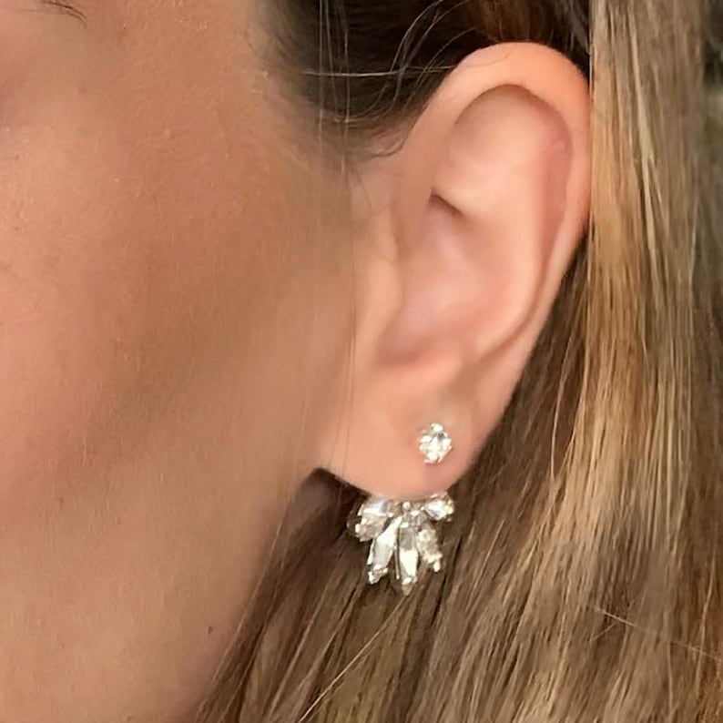 May include: A pair of silver earrings with clear crystal stones. The earrings are designed with a single stone in the front and a cluster of stones in the back.