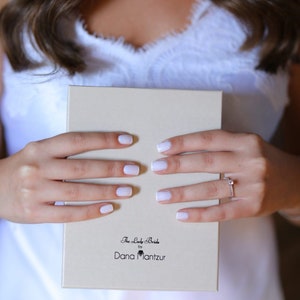 May include: A white box with the text "The Lady Bride by Dana Mantzur" on the front. The box is being held by a person with white nail polish.