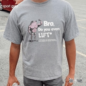 Puede incluir: Camiseta gris deportiva con una rana de dibujos animados flexionando su bíceps. El texto dice "Bro, Do you even LIFT?" con la frase "Lift others up. Show empathy. Hear people out. Choose kindness." debajo. La camiseta es de Funky Apparel.