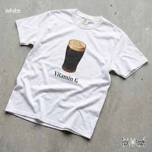 以下が含まれることがあります： 白いTシャツ。上面にクローバーのマークが付いた、濃いスタウトビールのグラフィック。ビール画像の下には「Vitamin G」の文字がプリントされています。このシャツは半袖で、クルーネックです。