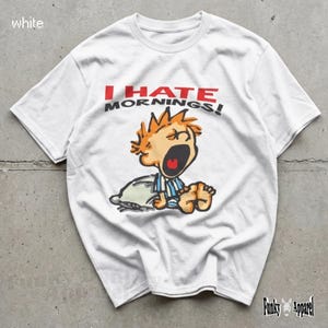 May include: White t-shirt with the text "I HATE MORNINGS!" in red and black. The shirt features a cartoon illustration of a yawning person in pyjamas. The shirt is from Funky Apparel.