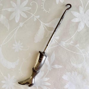 May include: A vintage silver-toned boot hook with a long, slender handle and a curved hook at the top. The boot hook features a detailed boot-shaped handle, showcasing a classic design. The metal has a slightly aged appearance.