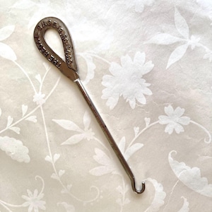 May include: A vintage metal crochet hook with a loop handle. The handle is stamped with the text "THE RECAT" and "SHOE E. 18.56-34.00". The hook has a long, slender shaft and a curved end. The background is a floral pattern.
