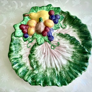 May include: A ceramic serving dish shaped like a green cabbage leaf with a raised center section. The center section is decorated with a cluster of fruit, including pears, grapes, and figs, painted in realistic colors.