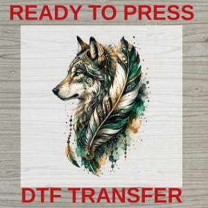 May include: A DTF transfer sheet featuring a wolf profile illustration with intricate green and gold patterns, accented by a large feather and hanging beads. The text READY TO PRESS and DTF TRANSFER is printed in red at the top and bottom of the image.