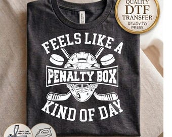 May include: A dark charcoal gray t-shirt featuring a white graphic print of a hockey goalie mask, two crossed hockey sticks, and two pucks. The text reads FEELS LIKE A PENALTY BOX KIND OF DAY. A gold circular badge states HIGH QUALITY DTF TRANSFER.