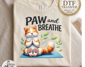 May include: A cream-colored t-shirt featuring a graphic of an orange and white cat sitting in a meditative pose on a blue mat. The text above the cat reads PAW and BREATHE. The design includes green leaves and small yellow dots.