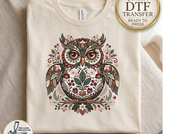 May include: A cream-colored t-shirt featuring a central graphic of an owl decorated with intricate floral and leaf patterns in shades of green, red, and brown. The owl has large eyes and is perched on a branch. A circular badge reads HIGH QUALITY DTF TRANSFER.