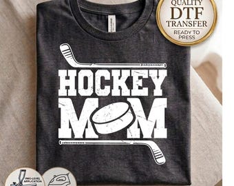 May include: A dark charcoal gray t-shirt featuring a white graphic print with the text HOCKEY MOM, a hockey puck replacing the letter O, and two hockey sticks framing the text. A gold circular badge reads HIGH QUALITY DTF TRANSFER.