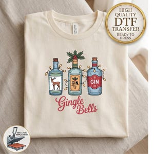 May include: A cream-colored t-shirt featuring a graphic of three glass bottles wrapped in string lights. The bottles are labeled GIN and decorated with a deer and holly. Below the bottles, the red text reads Gingle Bells. A gold seal reads High Quality DTF Transfer Ready to Press.