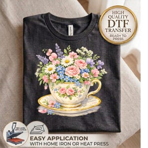 May include: A dark charcoal gray t-shirt featuring a graphic of a vintage-style teacup and saucer filled with a colorful bouquet of pink roses, white daisies, blue forget-me-nots, and purple lavender sprigs, with a gold-rimmed cup design.