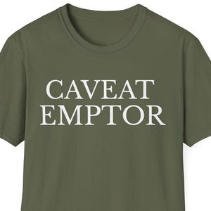 May include: Olive green t-shirt with the words "CAVEAT EMPTOR" printed in white block letters. The shirt has short sleeves and a crew neck. The text is centered on the front of the shirt.