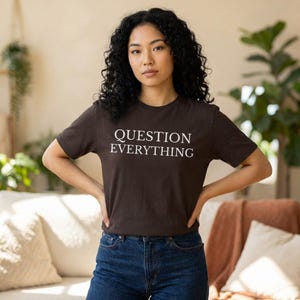Puede incluir: Una camiseta marrón con la frase "QUESTION EVERYTHING" en blanco. La persona lleva vaqueros azules. El fondo muestra un sofá crema y plantas de interior.