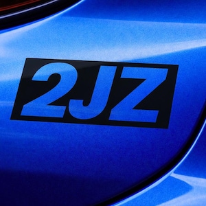 Può includere: Primo piano della carrozzeria blu di un'auto con un adesivo rettangolare nero. L'adesivo presenta il testo blu "2JZ". La vernice dell'auto ha una lucentezza metallica.