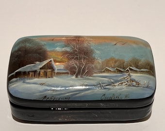 May include: A rectangular, black lacquered box with a winter landscape painting on the lid. The painting depicts a snow-covered village scene with a sunset sky. The box has text in a script font.