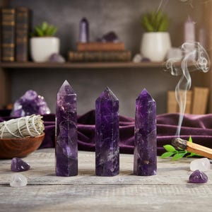 以下が含まれることがあります： 木製の表面に、自然な縞模様のある濃い紫色の3つのアメジストクリスタルタワーが配置されています。クリスタルが焦点で、植物、本、その他のクリスタルが置かれた棚が背景にあります。燃えているお香から煙が立ち上っています。