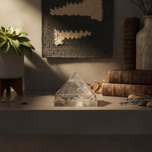 May include: A clear quartz crystal pyramid sits on a gray surface. The pyramid is the focal point, with a textured wall hanging and vintage books in the background. A potted succulent and decorative items complete the scene.