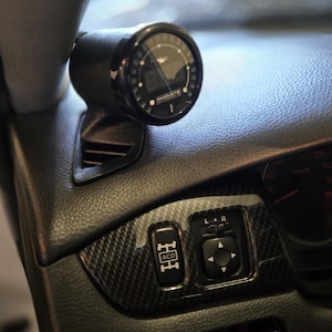 May include: Close-up of a car dashboard featuring a black gauge with the text "INNOVATE". The dashboard has carbon fiber accents and various buttons, including one labeled "ACD". The speedometer is visible in the background.
