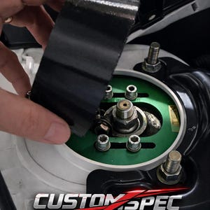 Puede incluir: Primer plano de un componente de suspensión de coche con cinta adhesiva negra que se está aplicando. El componente tiene un diseño verde y plateado con múltiples pernos. El texto "CUSTOM SPEC" es visible.