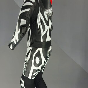 May include: Black and white motorcycle racing suit with a bold zebra-stripe pattern. The suit has a high collar, reinforced shoulders, and knee protection. The brand logo is visible on the chest. A red triangle is on the back.