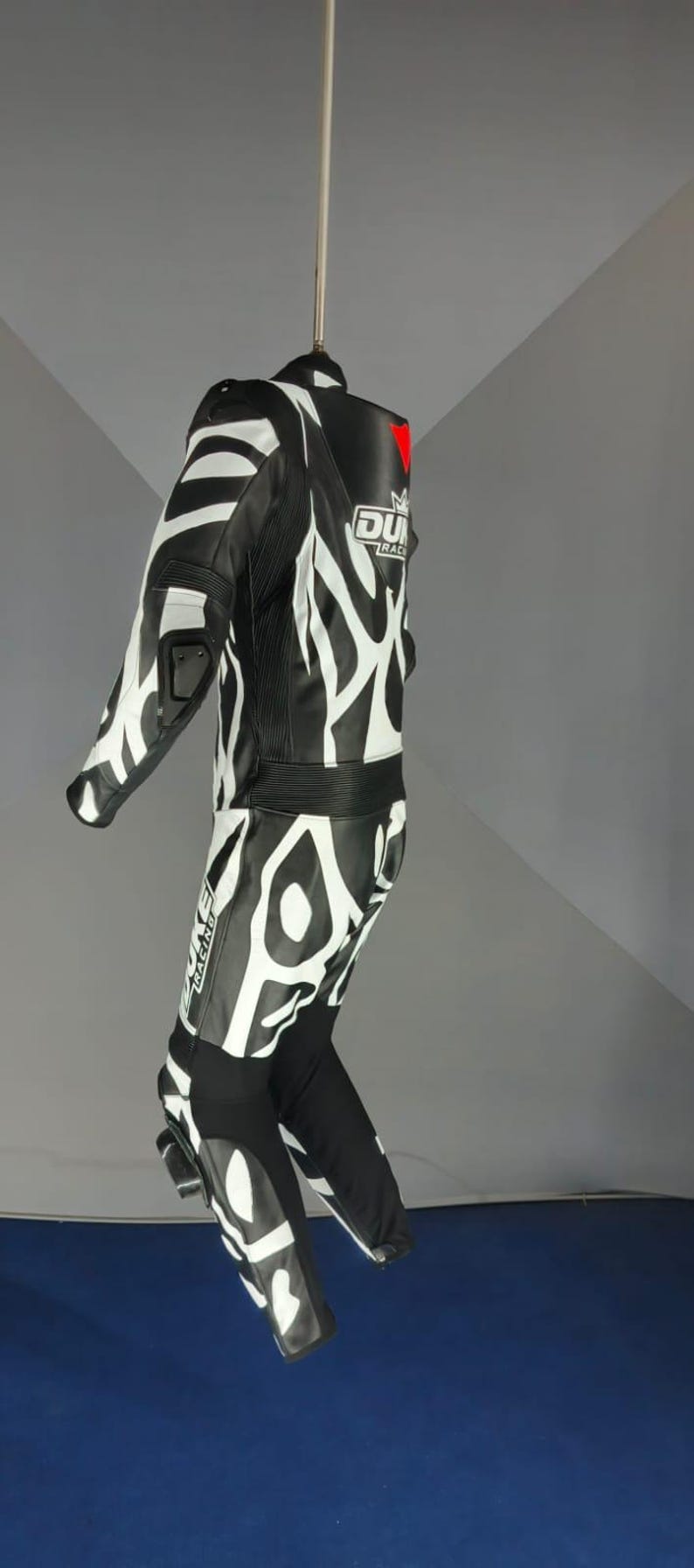 May include: Black and white motorcycle racing suit with a bold zebra-stripe pattern. The suit has a high collar, reinforced shoulders, and knee protection. The brand logo is visible on the chest. A red triangle is on the back.