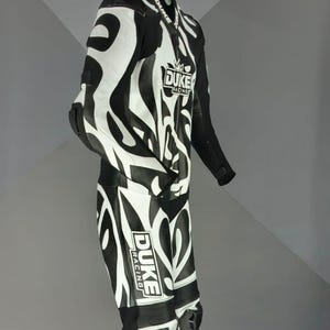 May include: Black and white motorcycle racing suit with an abstract pattern. The suit displays the "DUKE RACING" logo and is designed for protection. The suit is suspended from a hanger.