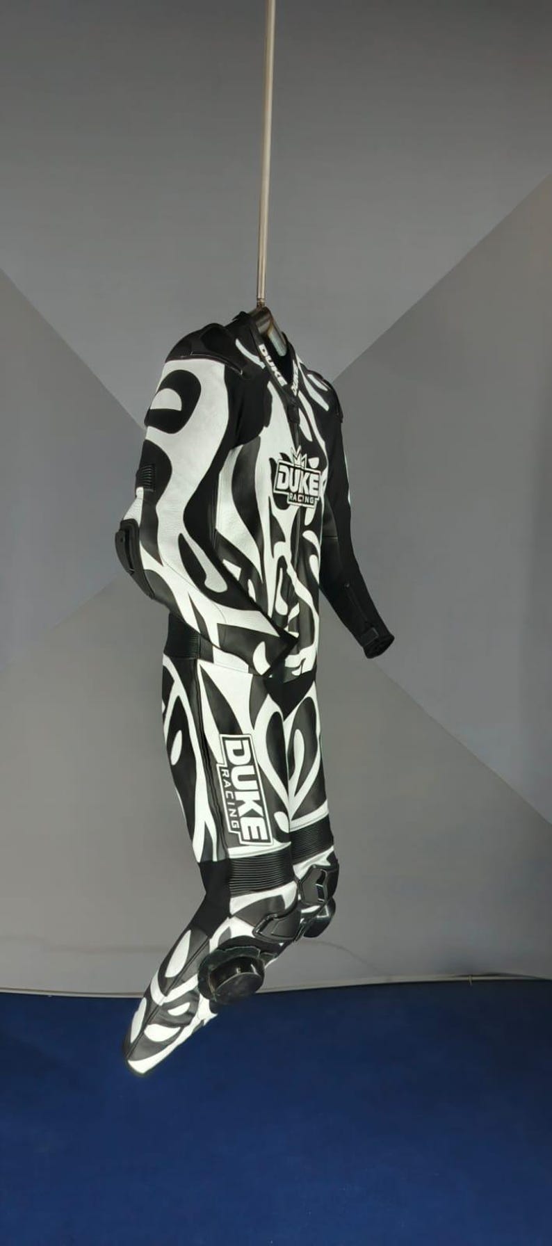 May include: Black and white motorcycle racing suit with an abstract pattern. The suit displays the "DUKE RACING" logo and is designed for protection. The suit is suspended from a hanger.