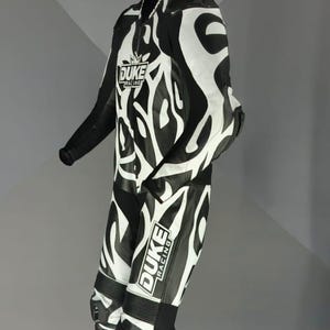 May include: Black and white motorcycle racing suit with an abstract design. The suit displays the "DUKE RACING" logo on the chest and leg. The suit is suspended from a hanger.