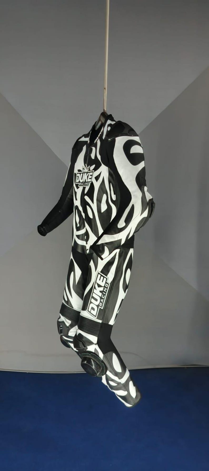 May include: Black and white motorcycle racing suit with an abstract design. The suit displays the "DUKE RACING" logo on the chest and leg. The suit is suspended from a hanger.