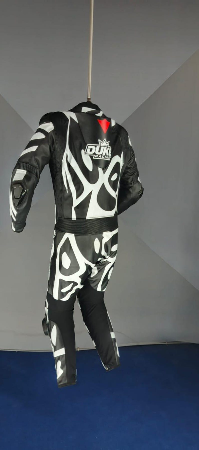 May include: Black and white motorcycle racing suit with an abstract pattern. The suit has the "DUKE RACING" logo and a red accent. Includes protective padding on the shoulders, elbows, and knees.