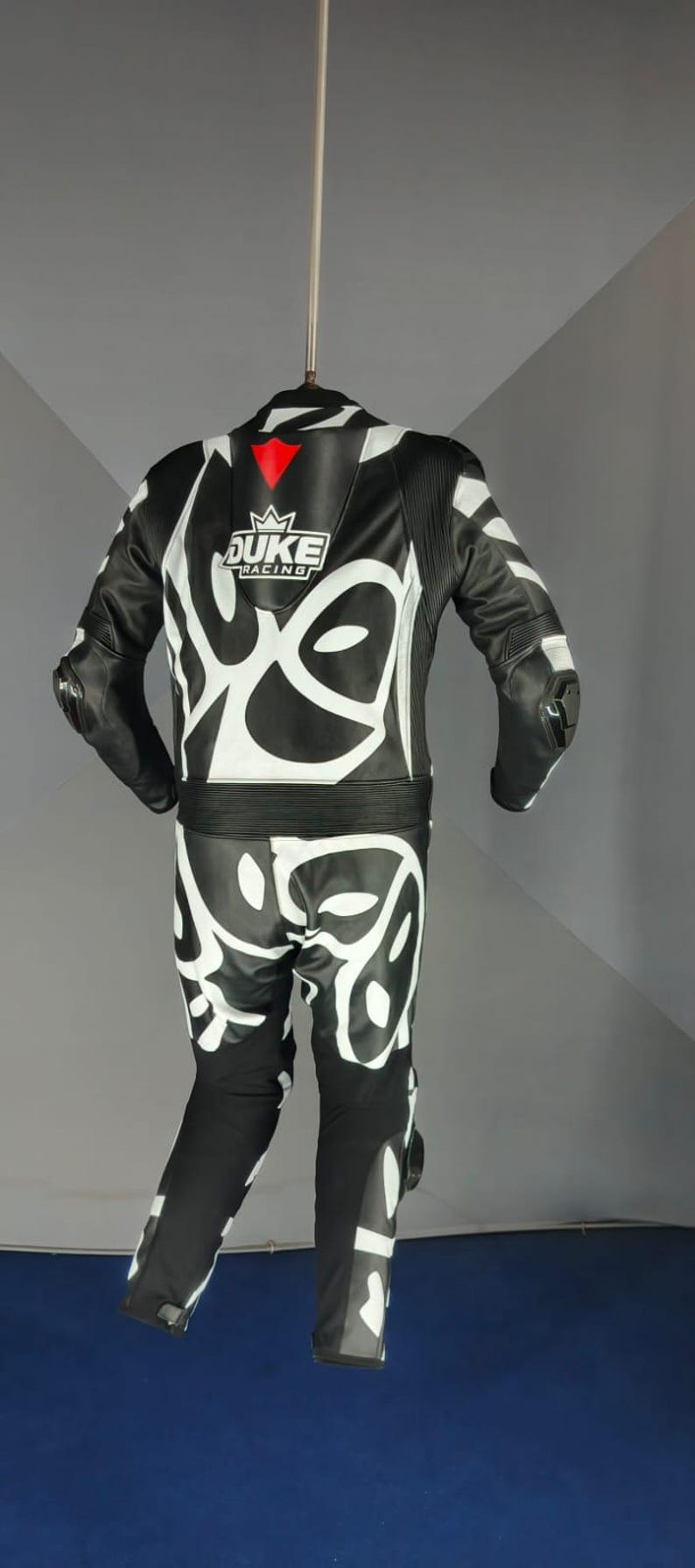 May include: Black and white motorcycle racing suit with a striking graphic pattern. The suit displays the "DUKE RACING" logo in white with red details. Designed for safety and performance on the track.