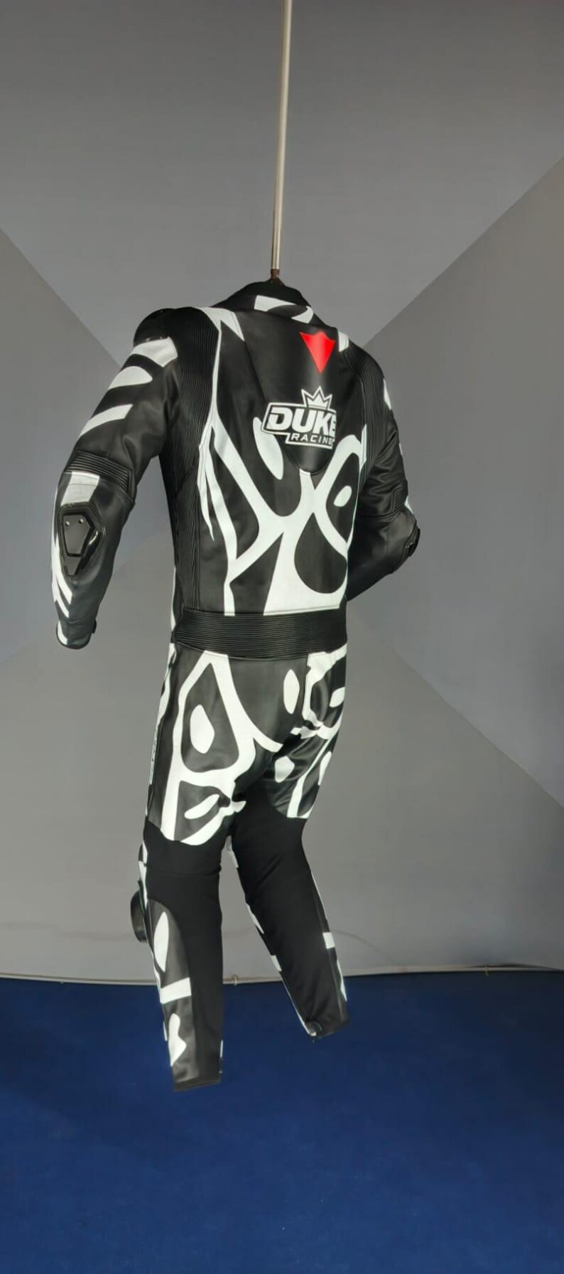 May include: Black and white motorcycle racing suit with the text "DUKE RACING". The suit features white abstract designs. The suit is suspended from a hanger.