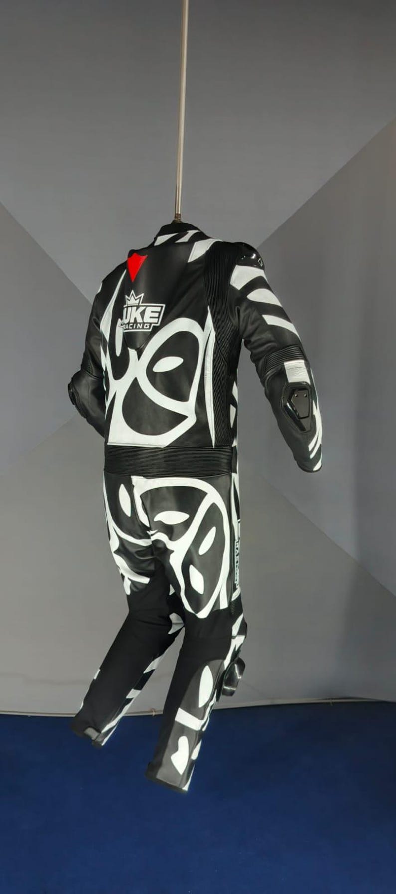 May include: Black and white motorcycle racing suit with a striking graphic pattern. The suit displays the "JUKE RACING" logo and red detailing. Constructed from leather, it includes protective padding on the shoulders, elbows, and knees.