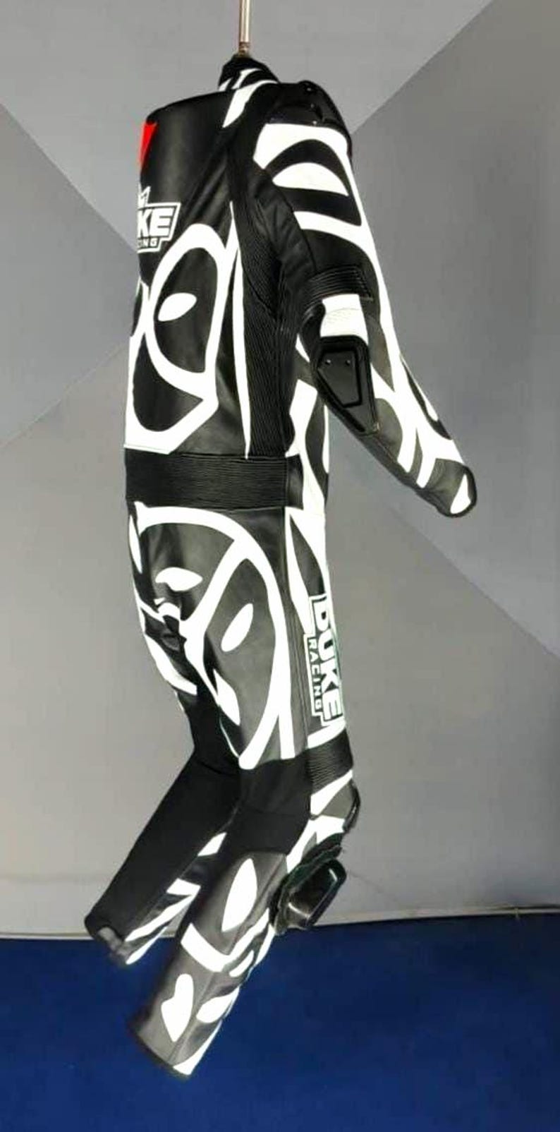 May include: Black and white motorcycle racing suit with a striking abstract pattern. The suit displays the "DUKE RACING" logo and protective padding. The suit is suspended from a hanger.