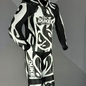 May include: Black and white motorcycle racing suit with an abstract pattern. The suit displays the "DUKE RACING" logo. The suit is suspended from a hanger, showcasing its design and features.