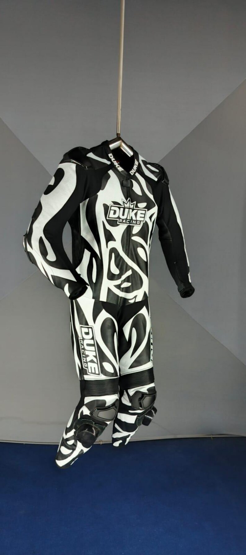 May include: Black and white motorcycle racing suit with an abstract pattern. The suit displays the "DUKE RACING" logo. The suit is suspended from a hanger, showcasing its design and features.