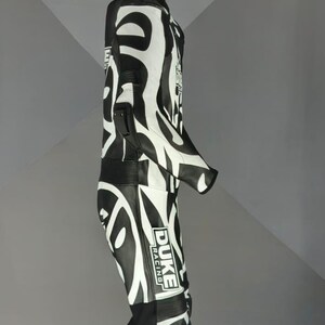 May include: Black and white racing suit with a striking zebra pattern. The suit has the "DUKE RACING" logo on the leg. The suit is suspended from a metal rod.