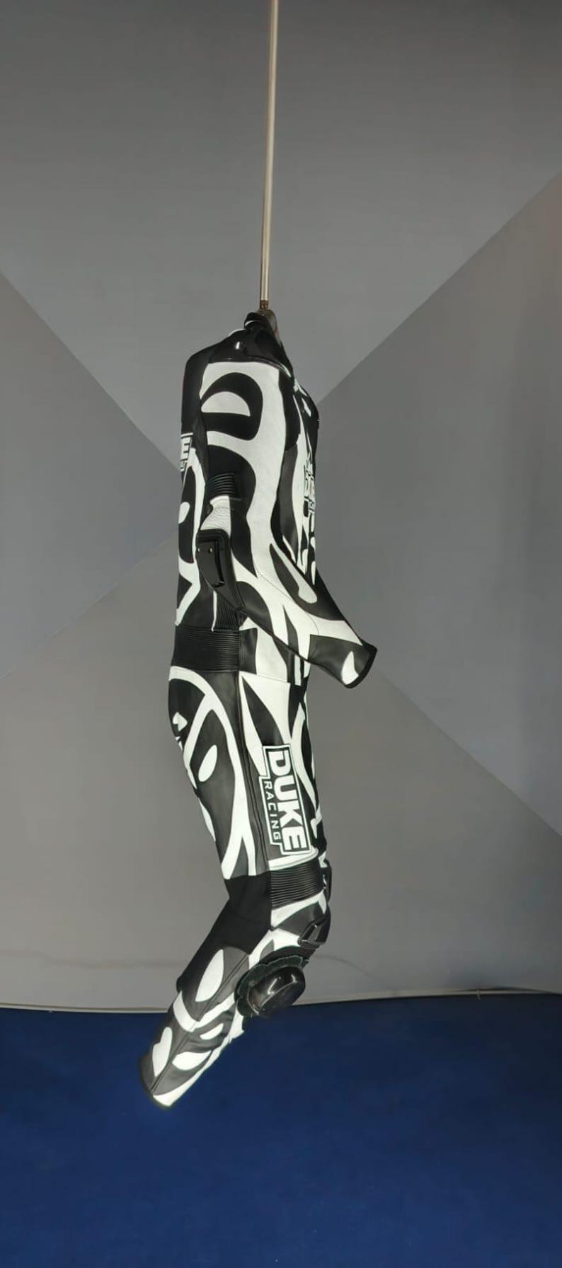 May include: Black and white racing suit with a striking zebra pattern. The suit has the "DUKE RACING" logo on the leg. The suit is suspended from a metal rod.
