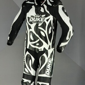 May include: Black and white motorcycle racing suit with an abstract design. The suit features the "DUKE Racing" logo on the chest and leg. The suit is hanging from a hanger.