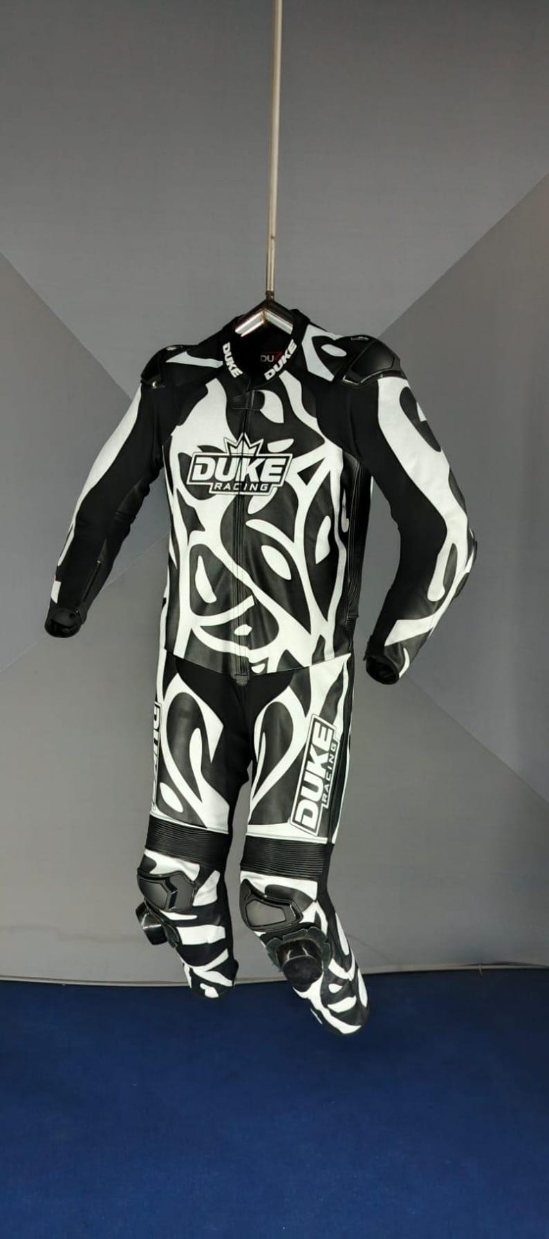 May include: Black and white motorcycle racing suit with an abstract design. The suit features the "DUKE Racing" logo on the chest and leg. The suit is hanging from a hanger.
