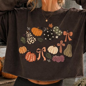 Peut inclure: Un sweat-shirt à manches longues marron foncé avec une impression graphique de diverses citrouilles, nœuds et feuilles. Les citrouilles sont décorées de motifs incluant des taches de léopard, des fleurs blanches et des rayures, avec une croix.
