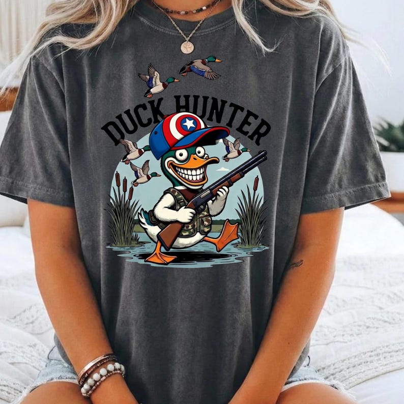 Op de afbeelding: Donkergrijs T-shirt met een cartoon eendenjager met een geweer, een camouflagevest en een rood, wit en blauwe hoed. De tekst "DUCK HUNTER" staat boven de eend. Eenden vliegen op de achtergrond.