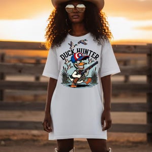 Op de afbeelding: Wit T-shirt met een cartoon eendenjager ontwerp. Het ontwerp toont een eend met een geweer, een hoed met een ster en de tekst "DUCK HUNTER". Eenden vliegen op de achtergrond. Het T-shirt wordt door een persoon gedragen.