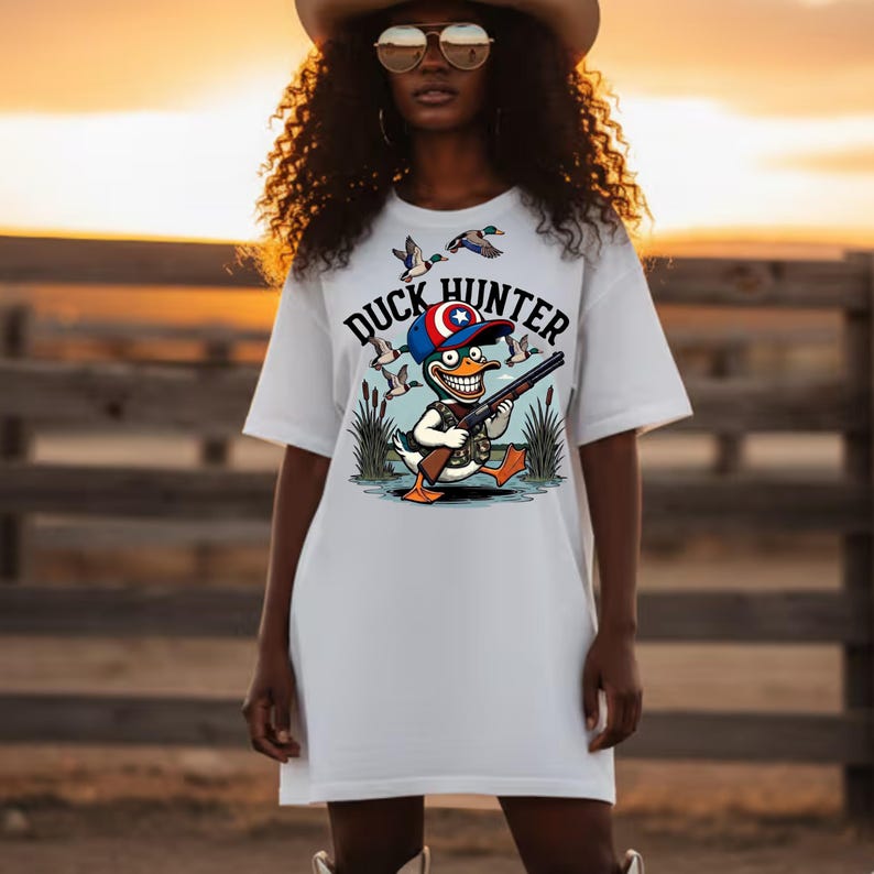 Op de afbeelding: Wit T-shirt met een cartoon eendenjager ontwerp. Het ontwerp toont een eend met een geweer, een hoed met een ster en de tekst "DUCK HUNTER". Eenden vliegen op de achtergrond. Het T-shirt wordt door een persoon gedragen.