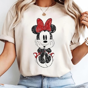 May include: A beige t-shirt featuring a distressed graphic of Minnie Mouse. The cartoon character is depicted in black and white with a red bow and shoes. The design is centered on the shirt, creating a vintage, worn look.
