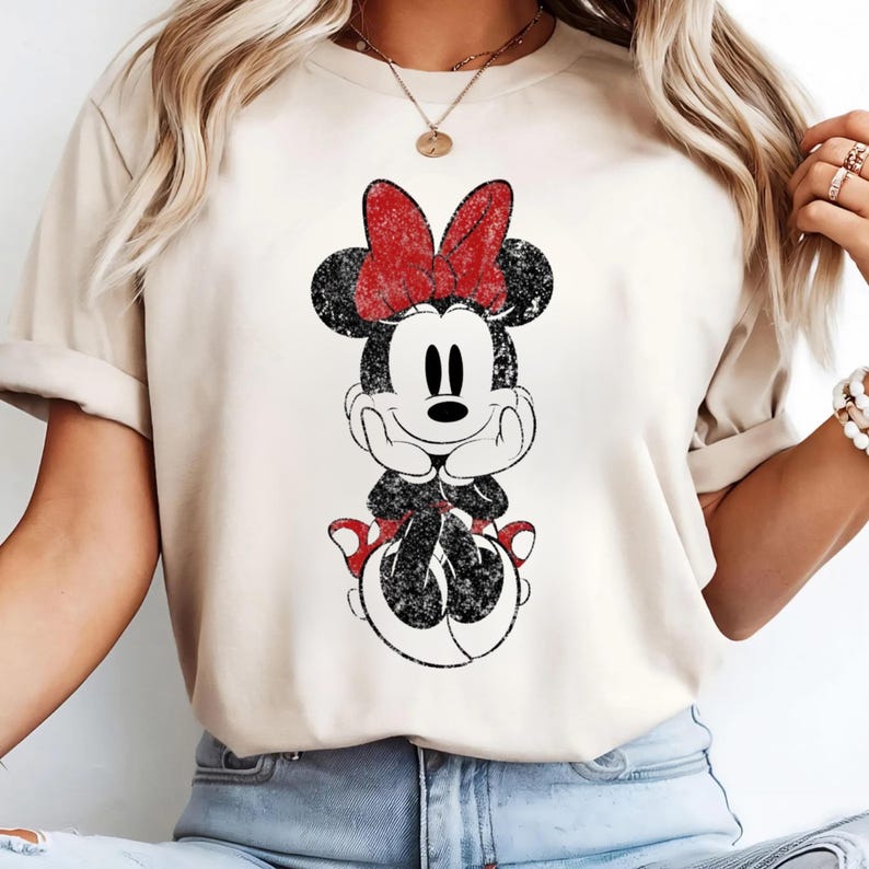 May include: A beige t-shirt featuring a distressed graphic of Minnie Mouse. The cartoon character is depicted in black and white with a red bow and shoes. The design is centered on the shirt, creating a vintage, worn look.