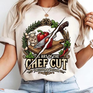 Peut inclure: Un t-shirt couleur crème avec un motif représentant un chaton portant une toque de chef, un poivron rouge sur une planche à découper en bois et un couteau. Le texte indique THE ART OF THE CHEF CUT PRECISION & PASSION, entouré d'herbes et de blé.