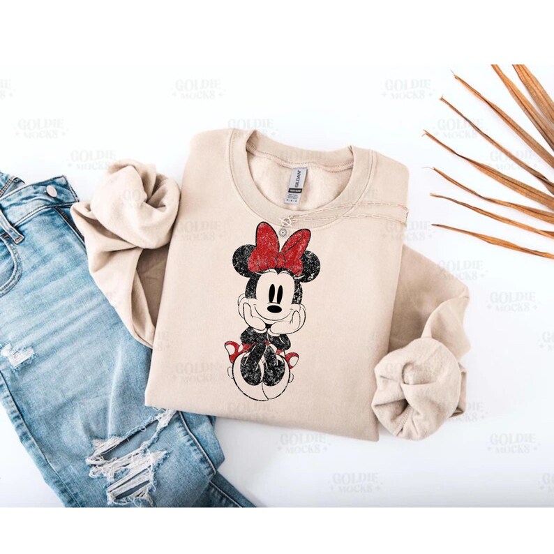 May include: Beige crewneck sweatshirt featuring a distressed graphic of Minnie Mouse with a red bow. The cartoon character is in a seated pose with her hands under her chin. The sweatshirt is paired with blue jeans.
