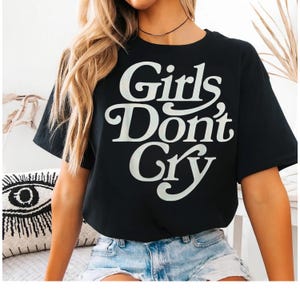 May include: Black t-shirt with the text "Girls Don't Cry" in a white, retro-style font. The shirt is worn with denim shorts. The overall aesthetic is casual and trendy.