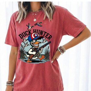 Op de afbeelding: Rood T-shirt met een cartoon eendenjager met een geweer, met een hoed met een ster. De tekst "DUCK HUNTER" staat boven de eend. Eenden vliegen op de achtergrond. Het shirt is effen rood.